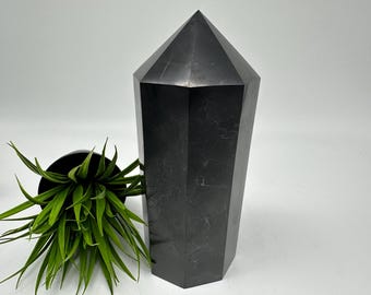 May include: A large, black obsidian crystal point, also known as a tower, with a smooth, polished surface. The crystal is sitting on a white surface.