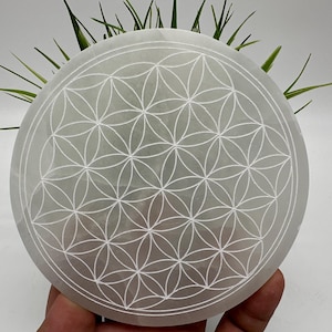 May include: A white selenite charging plate with a carved flower of life design. The plate is round and has a smooth, polished surface.