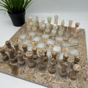 Peut inclure: Un jeu d'échecs artisanal avec un échiquier en pierre et des pièces d'échecs. Le plateau présente un motif en mosaïque avec des cases claires et foncées. Les pièces sont beiges et marron.