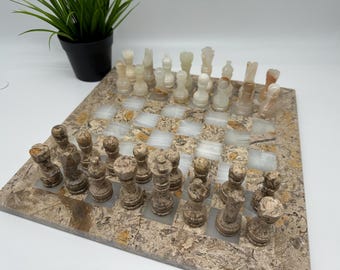 Puede incluir: Un juego de ajedrez artesanal con un tablero de piedra y piezas de ajedrez. El tablero presenta un patrón de mosaico con casillas claras y oscuras. Las piezas son de color beige y marrón.
