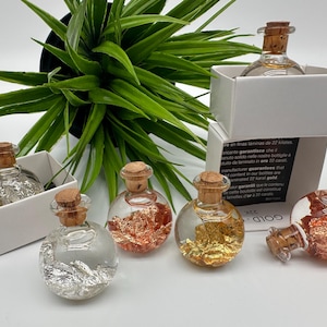 May include: Five small glass bottles with cork stoppers filled with clear liquid and gold, silver, and copper flakes. The bottles are arranged in a row on a white surface. The text "22K GOLD" is visible on the label of one of the bottles.