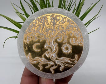 May include: A round, gray stone coaster with a gold-colored tree of life design. The tree features intricate details and is surrounded by a gold border. A crescent moon and sun are also included in the design. The coaster is held in a hand.