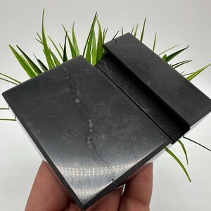 May include: A black marble phone stand with a slot for holding a phone. The stand is rectangular with a polished finish and a natural stone pattern. Green grass-like plants are behind the stand.