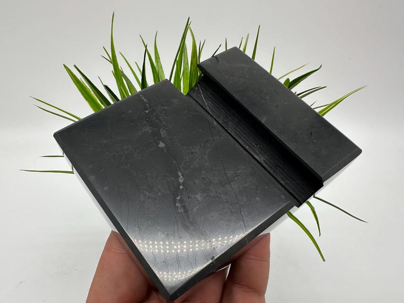 May include: A black marble phone stand with a slot for holding a phone. The stand is rectangular with a polished finish and a natural stone pattern. Green grass-like plants are behind the stand.