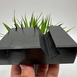 May include: A black, geometric phone stand with a slot for holding a phone. The stand is made of a solid, dark material and has a modern, minimalist design. Green plants are placed in the stand.