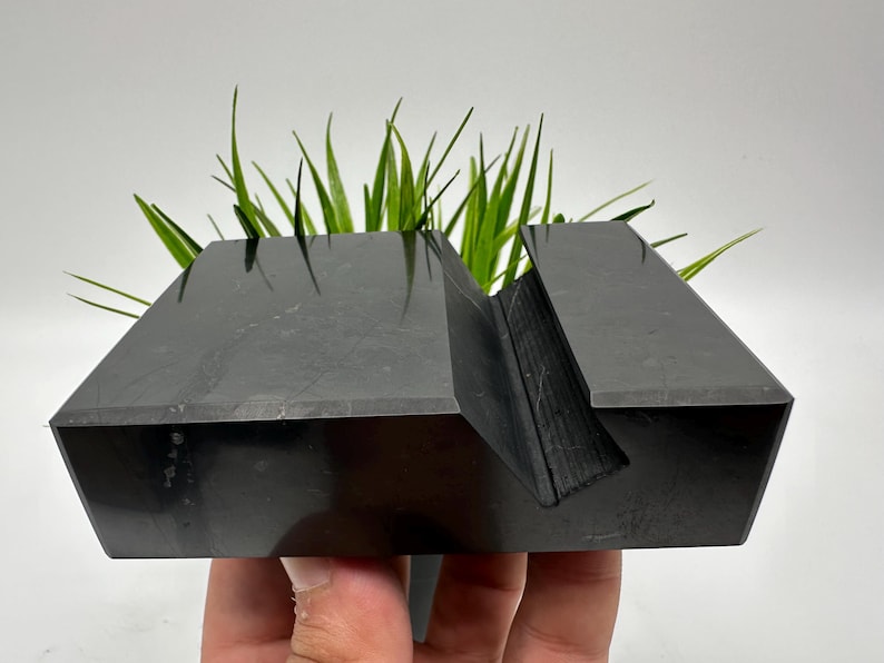 May include: A black, geometric phone stand with a slot for holding a phone. The stand is made of a solid, dark material and has a modern, minimalist design. Green plants are placed in the stand.
