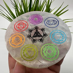May include: A white selenite chakra charging plate with seven colorful chakra symbols and a black and white image of a person meditating in the center.