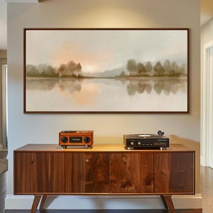 May include: A framed landscape painting depicts a serene lake scene with trees and a soft, muted color palette. The artwork hangs above a mid-century modern wooden cabinet, which holds a vintage-style radio and a record player.