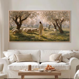 May include: A framed landscape painting depicts a figure in a flowing robe among a flock of sheep in a sunlit meadow. The scene is framed by blossoming trees, with a soft, natural color palette. The artwork is displayed above a white sofa.