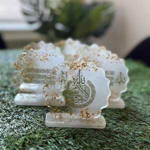 May include: Clear resin coasters with gold flecks and Arabic calligraphy. The coasters have a scalloped edge and a rectangular base. The calligraphy is in gold and green. The coasters are on a green surface.