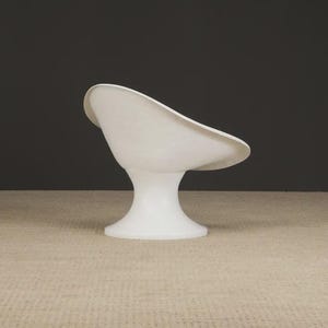 May include: A white, modern-style chair with a curved seat and a pedestal base. The chair is made of a smooth, glossy material and is set against a dark gray background. The chair's design is minimalist and sculptural.