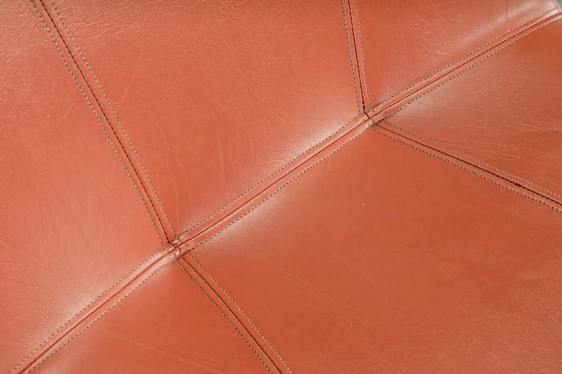 Pu&ograve; includere: Primo piano di pelle marrone-arancio cucita con filo scuro. La pelle &egrave; divisa in forme geometriche dalle cuciture, creando una superficie strutturata. Le cuciture sono visibili e aggiungono dettagli.