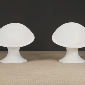 May include: Two white, mushroom-shaped chairs. Each chair has a rounded seat and a pedestal base. The chairs are made of a smooth, solid material and are set against a dark background. The chairs are a modern design.