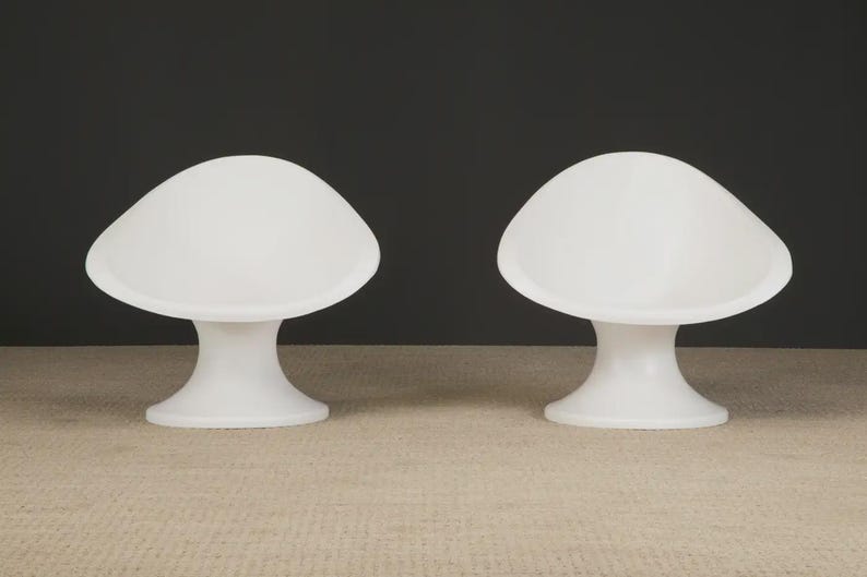 May include: Two white, mushroom-shaped chairs. Each chair has a rounded seat and a pedestal base. The chairs are made of a smooth, solid material and are set against a dark background. The chairs are a modern design.