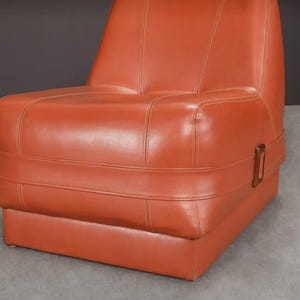 Pu&ograve; includere: Una poltrona in pelle arancione con base rettangolare e schienale alto. La sedia presenta dettagli cuciti e una fibbia in legno sul lato. La sedia &egrave; posta su uno sfondo grigio.
