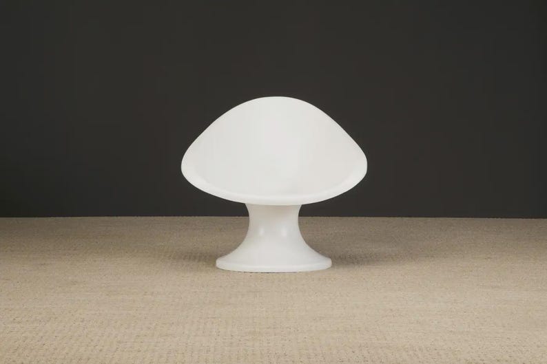 May include: A white, mushroom-shaped lamp with a rounded top and a flared base. The lamp is made of a smooth, opaque material and sits on a neutral-toned surface against a dark background. The lamp's height is approximately 10 inches.