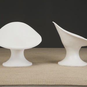 May include: Two white, modern-style chairs. One chair has a rounded, mushroom-like seat and a pedestal base. The other chair has a curved, open seat and a pedestal base. Both chairs are on a neutral-colored rug.