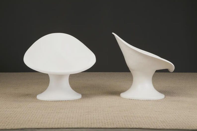 May include: Two white, modern-style chairs. One chair has a rounded, mushroom-like seat and a pedestal base. The other chair has a curved, open seat and a pedestal base. Both chairs are on a neutral-colored rug.