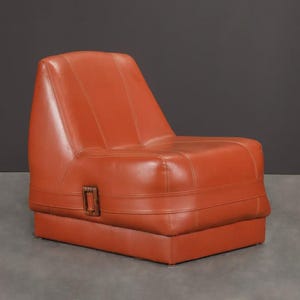 Pu&ograve; includere: Poltrona in pelle arancione con un profilo basso e uno schienale alto. La sedia ha una base rettangolare e una seduta con una leggera inclinazione. Un dettaglio con fibbia in metallo &egrave; visibile sul lato. La sedia &egrave; in stile moderno di met&agrave; secolo.