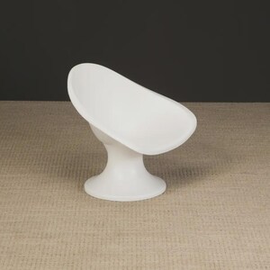 May include: A white, modern soap dish with a curved, oval-shaped bowl and a pedestal base. The dish is made of a smooth, solid material and is set against a neutral background. The design is minimalist and functional.