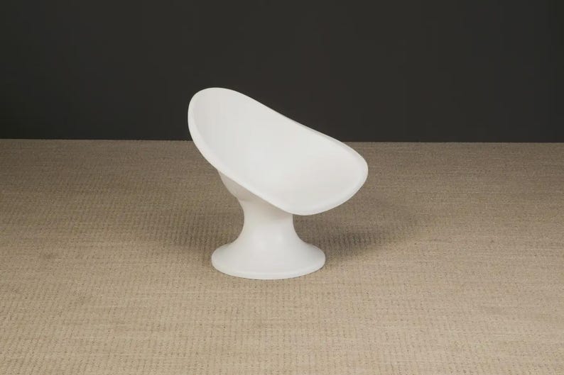 May include: A white, modern soap dish with a curved, oval-shaped bowl and a pedestal base. The dish is made of a smooth, solid material and is set against a neutral background. The design is minimalist and functional.