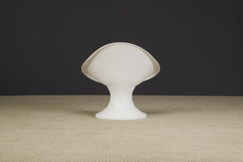 May include: A white, abstract, sculptural object with a flared top and a pedestal base. The object has a smooth, matte finish and is set against a dark background. The shape resembles a stylized shell or a mushroom.
