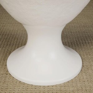 May include: A white, modern pedestal table base with a rounded top and a flared, hourglass-shaped stem. The base is made of a smooth, solid material and sits on a neutral-colored carpet.