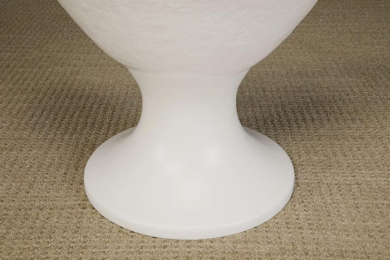 May include: A white, modern pedestal table base with a rounded top and a flared, hourglass-shaped stem. The base is made of a smooth, solid material and sits on a neutral-colored carpet.