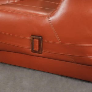 Pu&ograve; includere: Primo piano di un paio di poltrone in pelle arancione. Le sedie hanno una base rettangolare e uno schienale curvo. Un dettaglio con fibbia in legno &egrave; visibile sul lato della sedia. Le sedie sono rivestite in pelle liscia e lucida.