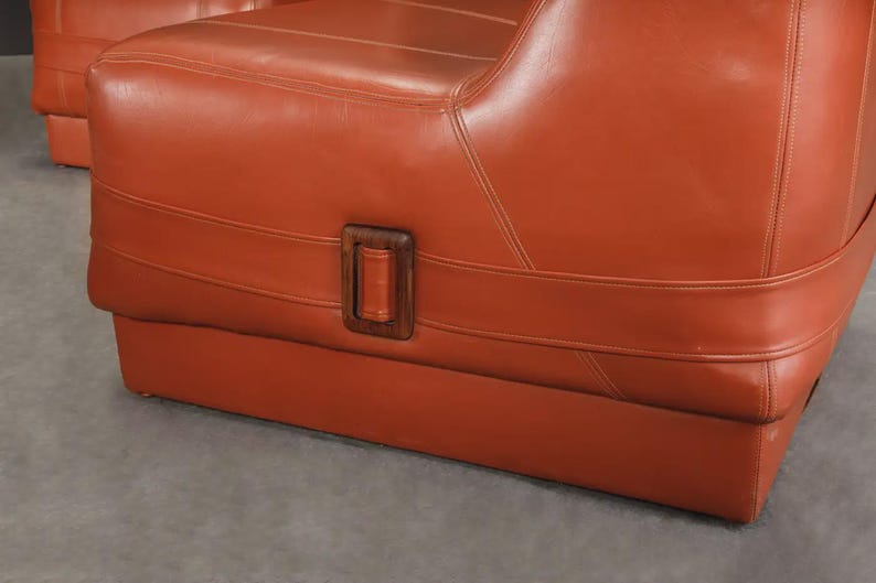 Pu&ograve; includere: Primo piano di un paio di poltrone in pelle arancione. Le sedie hanno una base rettangolare e uno schienale curvo. Un dettaglio con fibbia in legno &egrave; visibile sul lato della sedia. Le sedie sono rivestite in pelle liscia e lucida.