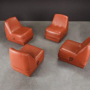 Pu&ograve; includere: Quattro poltrone in pelle arancione con un profilo basso e un design squadrato. Ogni sedia ha un piccolo dettaglio di fibbia rettangolare sul lato. Le sedie sono disposte su una superficie grigia.