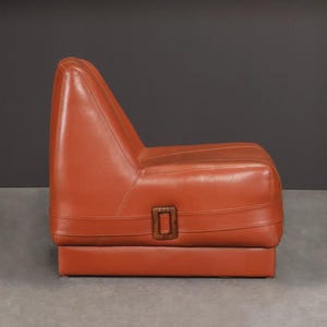 Pu&ograve; includere: Una poltrona in pelle arancione in stile moderno della met&agrave; del secolo, con schienale alto e base rettangolare. Ha un dettaglio con fibbia in legno sul lato. La poltrona &egrave; posta su uno sfondo grigio scuro.