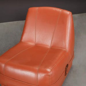 Pu&ograve; includere: Sedia in pelle arancione con schienale alto e base rettangolare. La sedia presenta dettagli di cucitura verticali e un accento in legno sul lato. La sedia &egrave; in stile moderno di met&agrave; secolo.