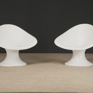 May include: Two white, modern-style chairs. Each chair has a rounded seat and a pedestal base. The chairs are made of a smooth, solid material and are set against a dark background. The chairs are placed on a light-colored surface.