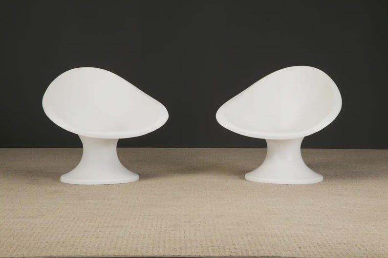 May include: Two white, modern-style chairs. Each chair has a rounded seat and a pedestal base. The chairs are made of a smooth, solid material and are set against a dark background. The chairs are placed on a light-colored surface.