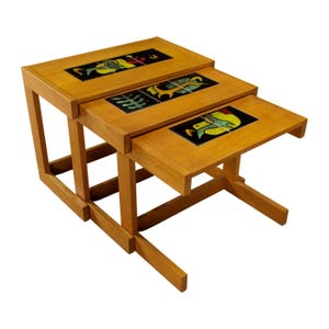 May include: Set of three nesting tables with a light wood finish. Each table has a rectangular top with a black tile inlay featuring colorful fish and plant designs. The tables are supported by sturdy, angled legs.