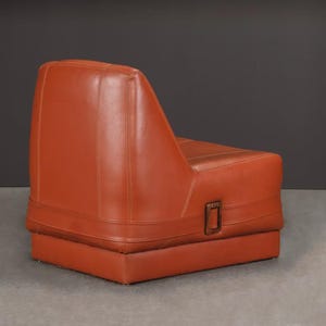 Pu&ograve; includere: Poltrona in pelle arancione con base rettangolare e schienale inclinato. La poltrona presenta cuciture decorative e una fibbia metallica. &Egrave; posta su uno sfondo grigio scuro. Design retr&ograve;.