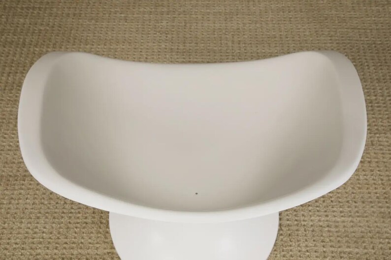 May include: A white, modern-style chair with a curved seat and a pedestal base. The chair has a smooth, matte finish and a minimalist design. The chair is likely made of plastic or a similar material.