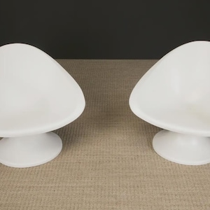 May include: Two white, modern-style chairs. Each chair has a rounded, triangular seat and a circular base. The chairs are made of a smooth, solid material and are set against a neutral background. The chairs are designed for indoor use.