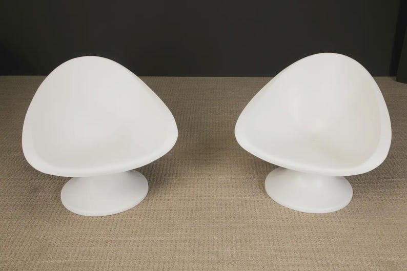 May include: Two white, modern-style chairs. Each chair has a rounded, triangular seat and a circular base. The chairs are made of a smooth, solid material and are set against a neutral background. The chairs are designed for indoor use.