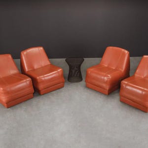 Pu&ograve; includere: Quattro sedie in pelle arancione-rossa e un tavolino marrone scuro. Le sedie sono disposte in gruppo, con il tavolino al centro. Le sedie hanno una forma squadrata e sono realizzate in un materiale liscio e lucido.