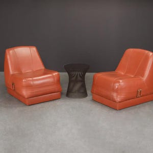 Pu&ograve; includere: Due poltrone in pelle arancione e un tavolino laterale marrone scuro. Le poltrone hanno un profilo basso e un design squadrato. Il tavolino &egrave; cilindrico con una trama intrecciata. L'arredamento &egrave; su sfondo grigio scuro.