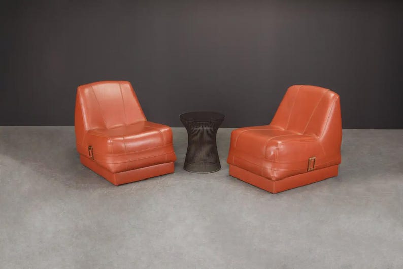 Pu&ograve; includere: Due poltrone in pelle arancione e un tavolino laterale marrone scuro. Le poltrone hanno un profilo basso e un design squadrato. Il tavolino &egrave; cilindrico con una trama intrecciata. L'arredamento &egrave; su sfondo grigio scuro.