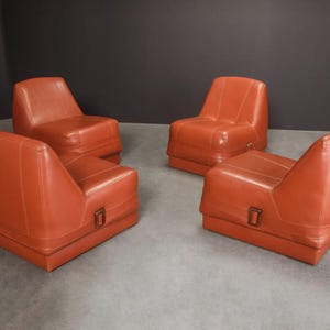 Pu&ograve; includere: Quattro poltrone in pelle arancione-rossa. Ogni sedia ha una base rettangolare con un dettaglio a fibbia e uno schienale curvo. Le sedie sono disposte in una stanza con pavimento grigio e parete grigio scuro.