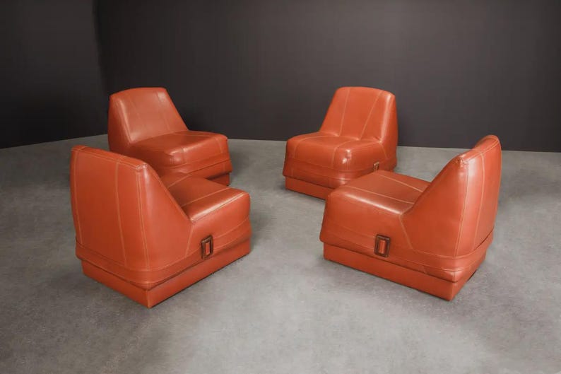 Pu&ograve; includere: Quattro poltrone in pelle arancione-rossa. Ogni sedia ha una base rettangolare con un dettaglio a fibbia e uno schienale curvo. Le sedie sono disposte in una stanza con pavimento grigio e parete grigio scuro.