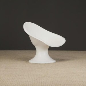 May include: A modern, white, sculptural chair with a curved seat and a pedestal base. The chair is made of a smooth, solid material and is set against a dark gray background. The chair's design is minimalist and elegant.