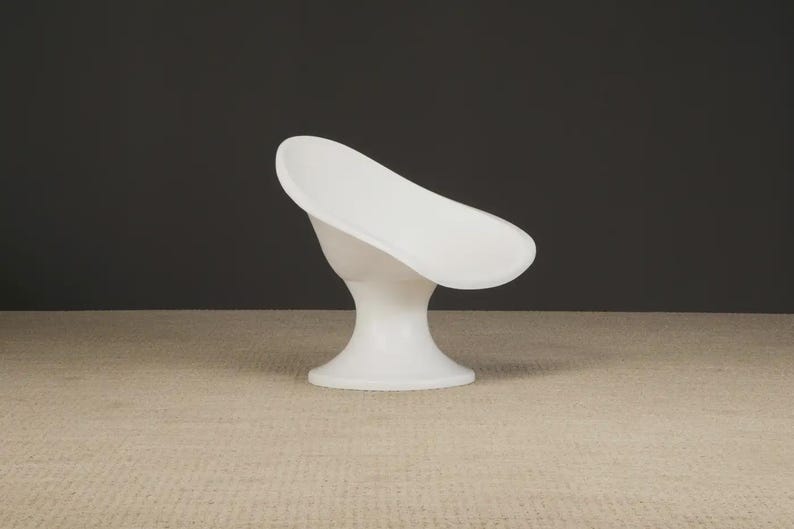 May include: A modern, white, sculptural chair with a curved seat and a pedestal base. The chair is made of a smooth, solid material and is set against a dark gray background. The chair's design is minimalist and elegant.
