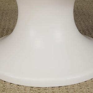 May include: A white, pedestal-style table base. The base has a rounded, flared design, with a wide, flat bottom. The surface is smooth and glossy. The table base is likely made of a durable material, such as plastic or composite.
