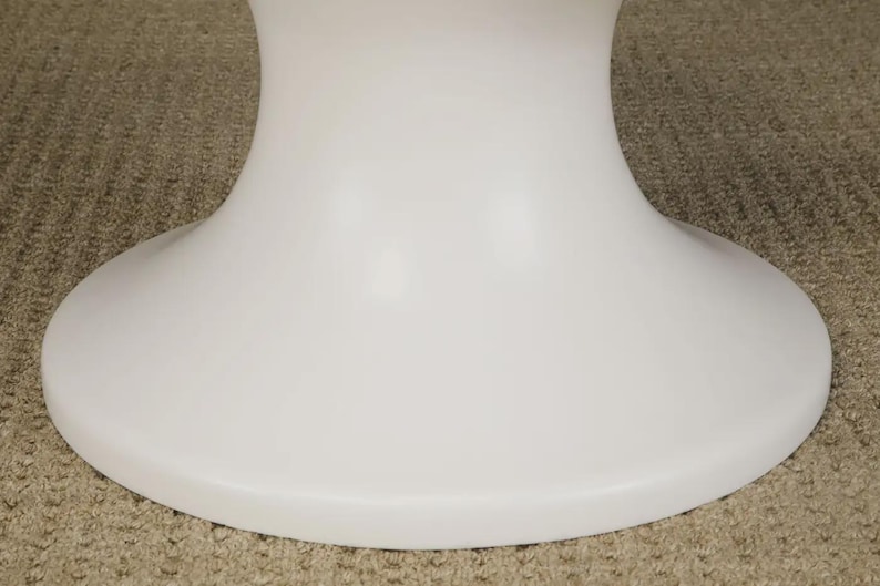 May include: A white, pedestal-style table base. The base has a rounded, flared design, with a wide, flat bottom. The surface is smooth and glossy. The table base is likely made of a durable material, such as plastic or composite.
