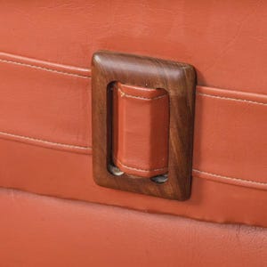 Pu&ograve; includere: Primo piano di una maniglia rettangolare in legno con impugnatura in pelle. La maniglia &egrave; posta su una superficie arancione strutturata con cuciture bianche. La cornice in legno &egrave; marrone scuro.